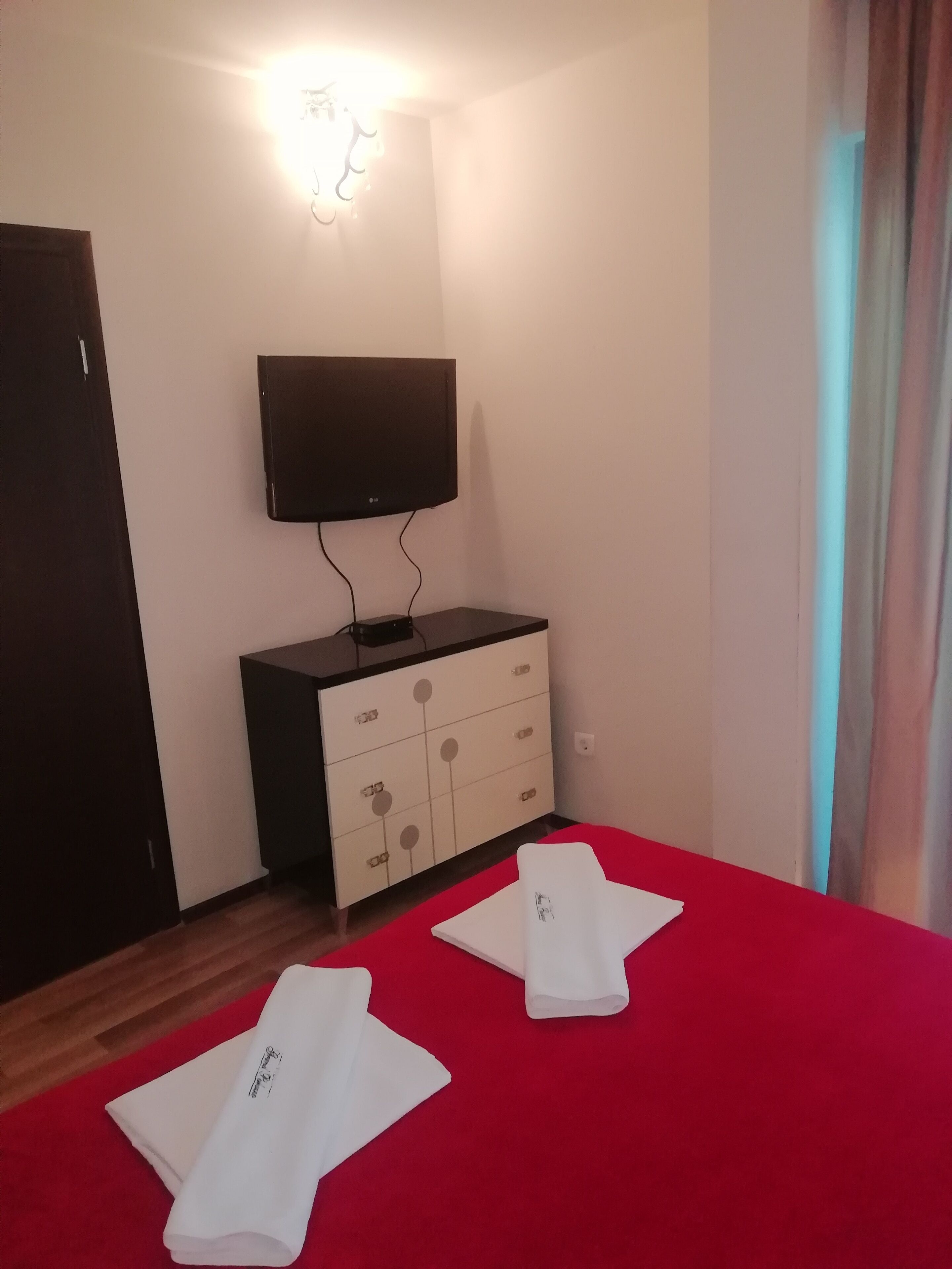 A hotel room with a flat-screen TV mounted on the wall, a wooden wardrobe, and a red bedspread.