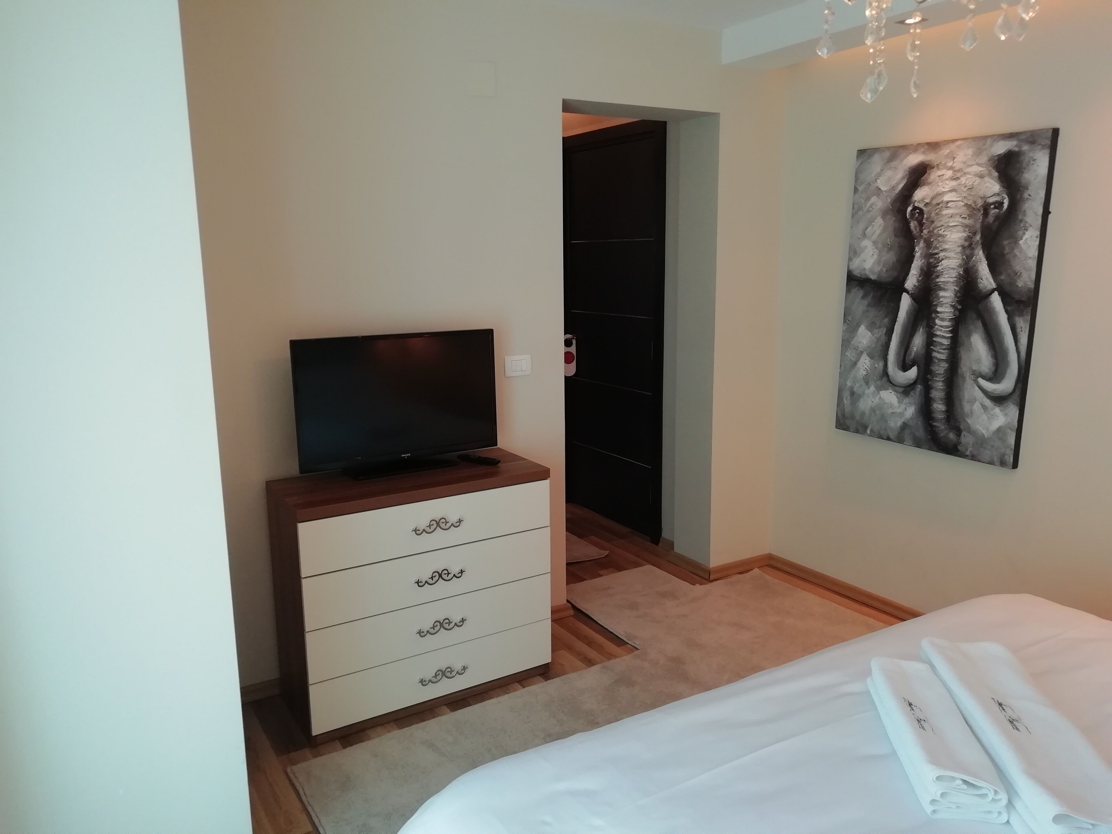A hotel room with a bed, a TV on a wooden cabinet, a painting of an elephant, and a door leading to another room.