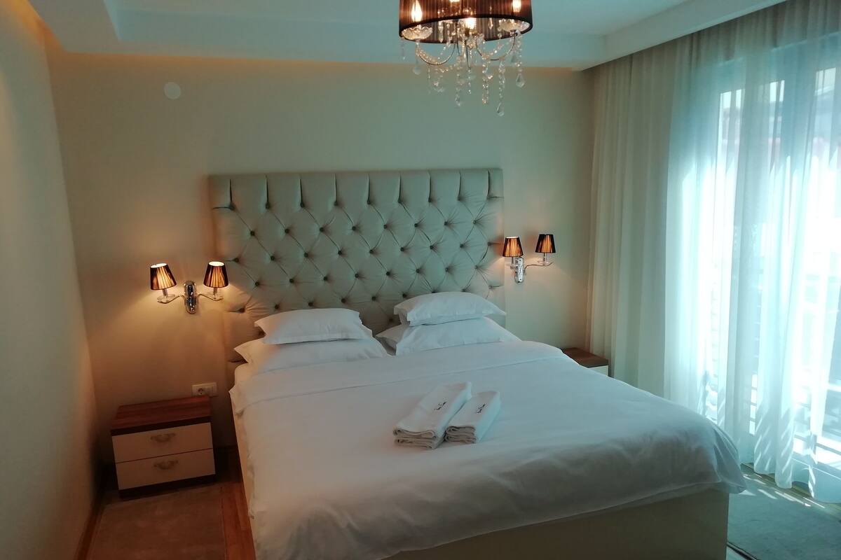 Una cama bien hecha con sábanas y almohadas blancas, un cabecero capitonado, lámparas de noche y una araña.