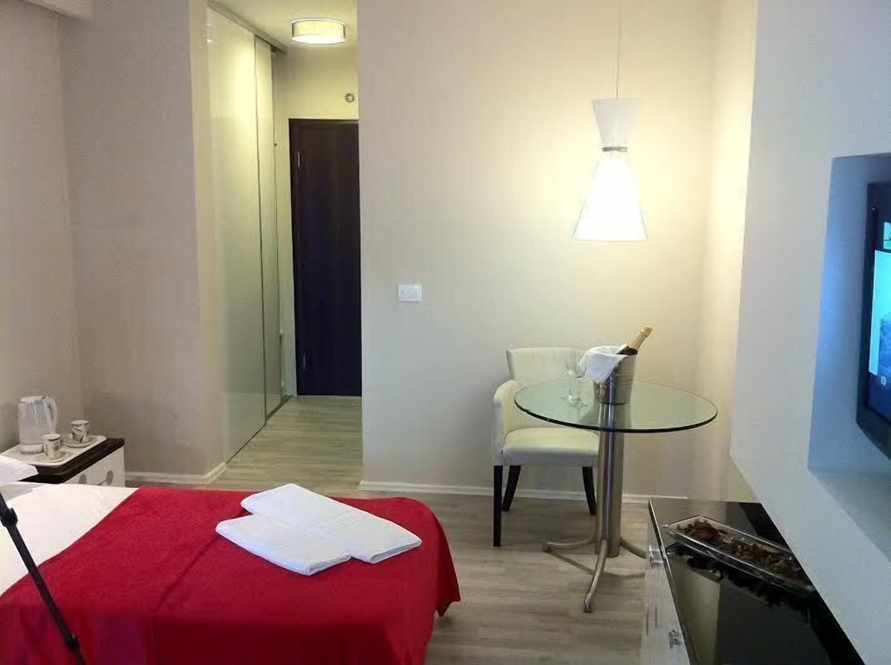 Habitación de hotel con una cama, una mesita con una botella y vasos, una silla y un televisor en la pared.