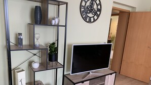 A modern living room with a TV on a wooden stand, a metal shelving unit with decorative items, and a large wall clock. - New! Stylish city apartment *Netflix,parking lot,close to BHF* (CB)