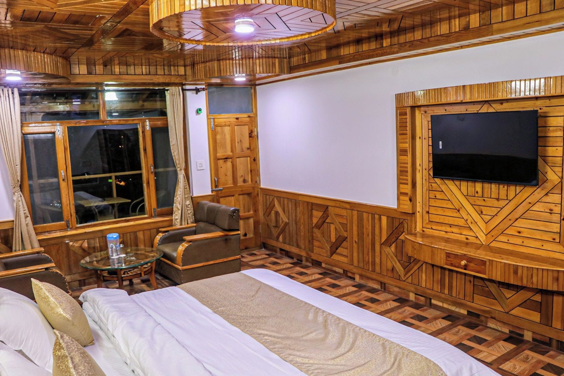 A wooden-themed room with a bed, seating area, and a television mounted on the wall.