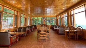 A wooden interior with a central dining table surrounded by chairs and benches. Large windows provide views of the outdoors.