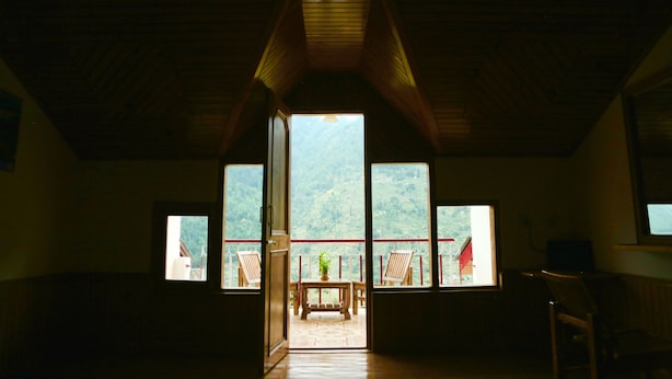 A room with a sloped ceiling and a large window revealing a balcony with a table and chairs.