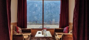 A room with two brown leather sofas, a small table with a vase and a bottle, and a large window with a view of a balcony and outdoor furniture. - Hotel Rishi Palace (Manali)