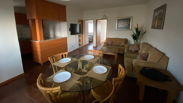 Uma sala de estar moderna com uma mesa de jantar de vidro, cadeiras de vime, um sofá bege e uma televisão na área da cozinha.
