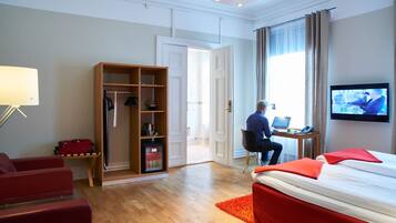 Een hotelkamer met een bed, een bureau met een laptop, een televisie, een kledingkast en een rood tapijt.