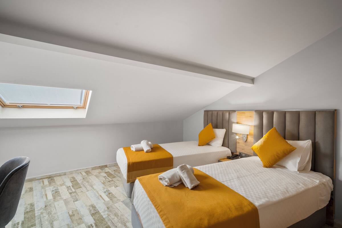 Quarto de hotel com duas camas, um claraboia, cabeceira moderna e uma pequena escrivaninha com abajur.