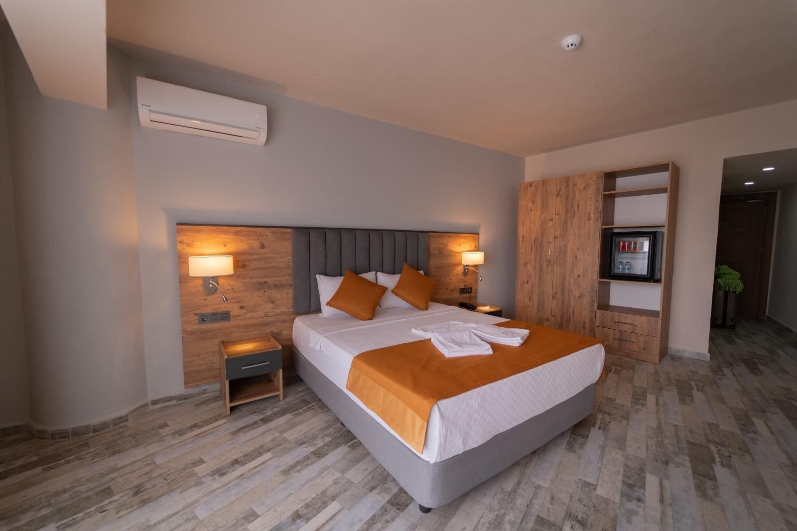 Una habitación de hotel moderna con una cama grande, cabecera de madera, dos mesitas de noche, un mueble empotrado para la televisión y un equipo de aire acondicionado.