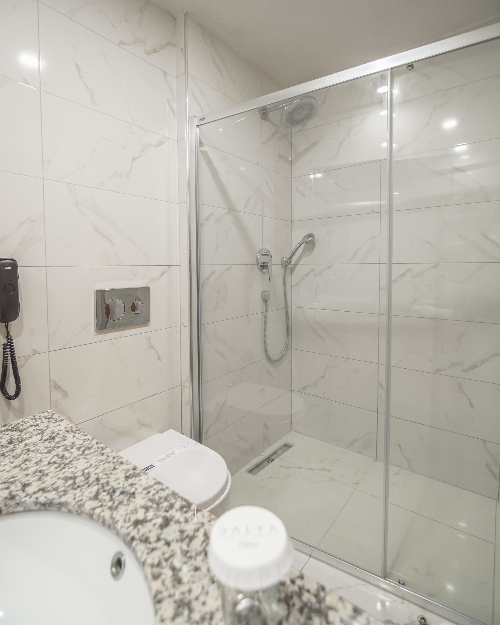 Ein Badezimmer mit einer Glaskabine zur Dusche, einer Wand mit Marmorplatten, einem Waschbecken mit Seifenspender und einer Toilette.