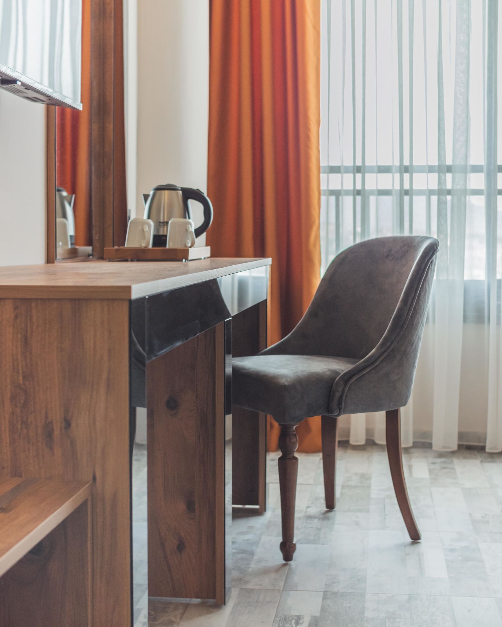 Ein modernes Zimmer mit einem Holzbüro, einem grau gepolsterten Sessel und einem großen Fenster mit orangefarbenen Vorhängen.