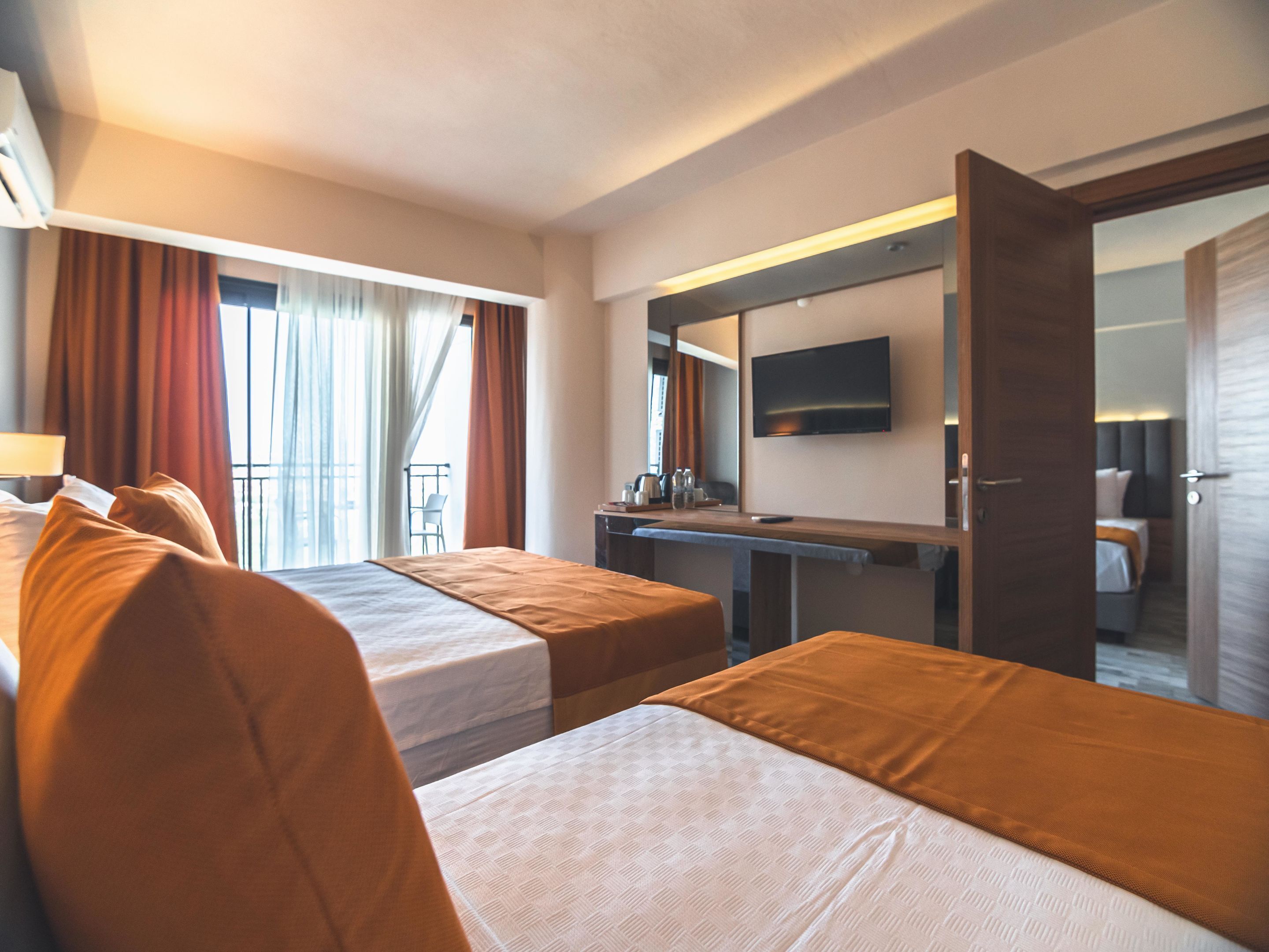 A hotel room with two beds, a desk, a TV, and a balcony with curtains.