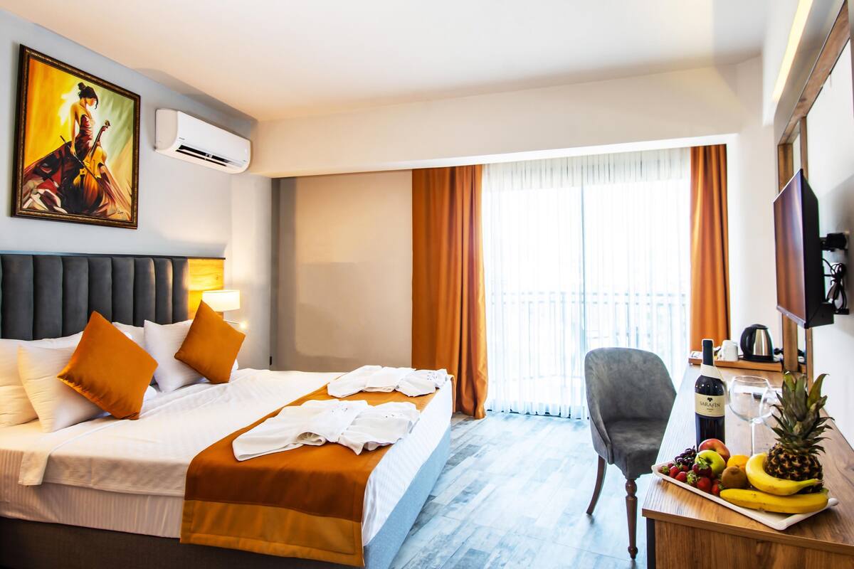 Quarto de hotel com uma cama, uma escrivaninha com frutas, uma cadeira e um quadro na parede.