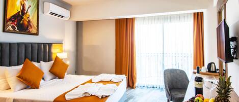 A hotel room with a bed, a desk with fruit, a chair, and a painting on the wall.