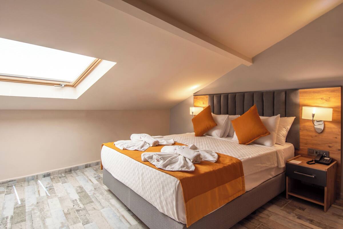 Quarto de hotel com claraboia, uma cama grande com roupa de cama laranja, cabeceira de madeira e mesa de cabeceira com abajur.