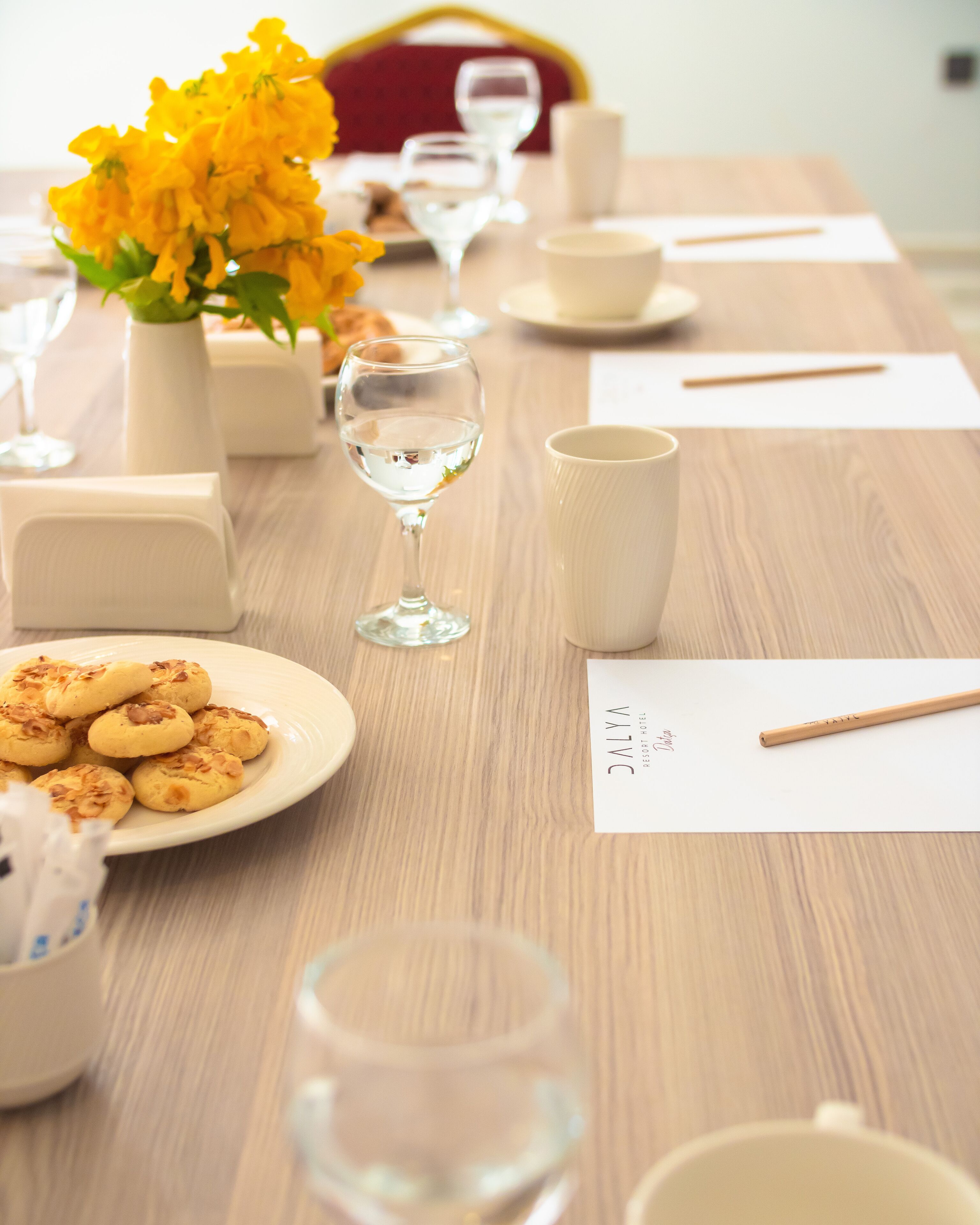 Mesa posta com um prato de biscoitos, um copo de água, um vaso de flores amarelas, uma caneca branca e uma colher de madeira em uma mesa de madeira.