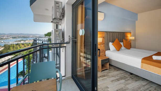 A balcony with a view of a body of water and distant landscape, a bed with pillows and a nightstand, and a table with chairs. - Dalya Resort Aqua & Spa Hotel (Datça)