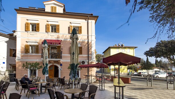 Un caffè all'aperto con sedie e tavoli in vimini, un edificio con il cartello 'Jurani', e un cielo azzurro limpido.