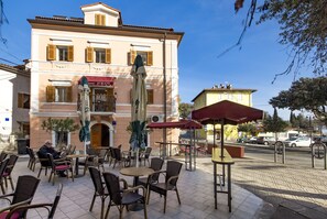 An outdoor cafe with wicker chairs and tables, a building with a 'Jurani' sign, and a clear blue sky. - Beautiful Apartment House On The Seaside (Opatija)