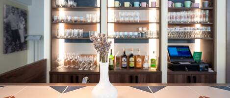 A bar area with shelves of glasses and bottles, a white vase with flowers, and a laptop on a counter.
