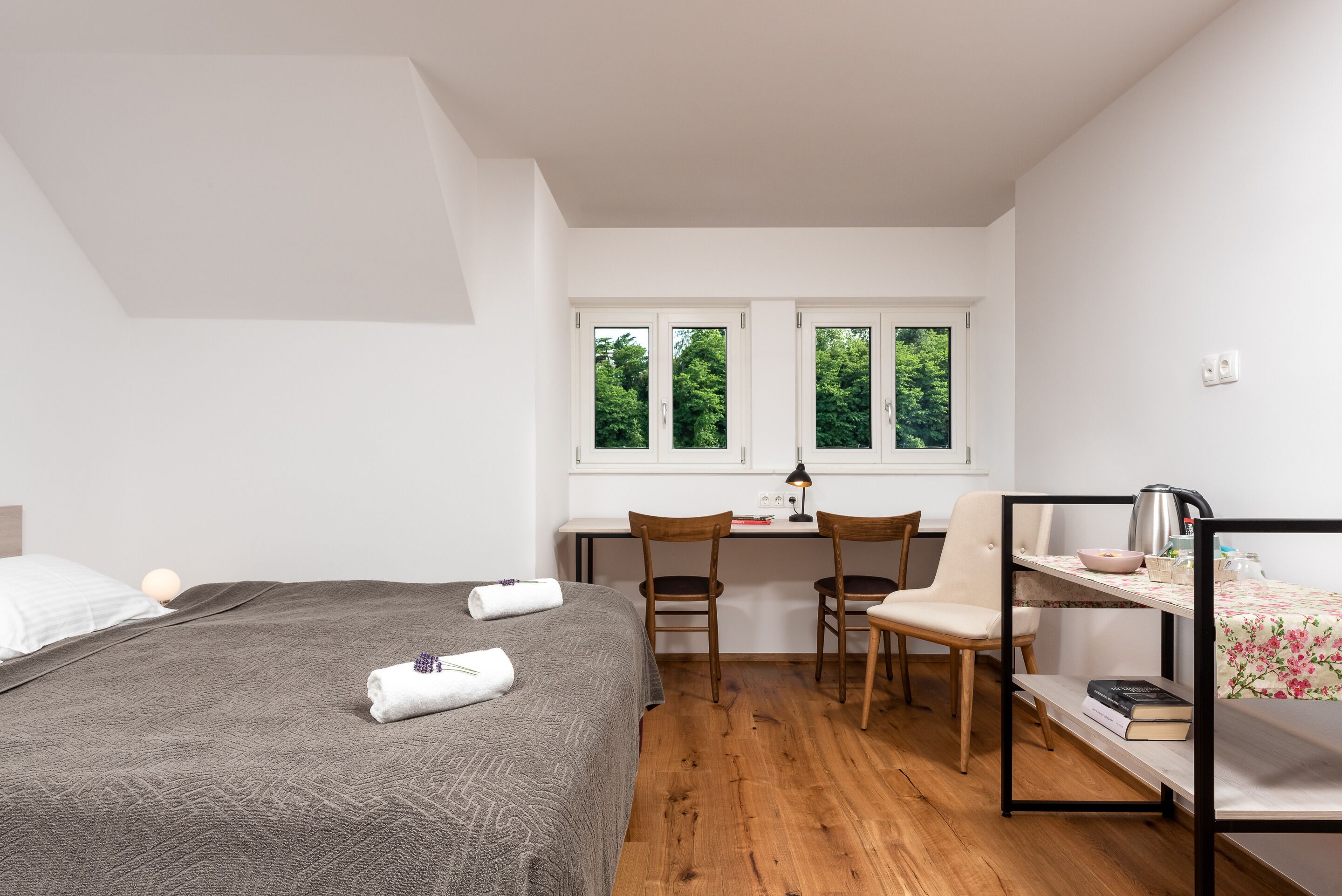 A bedroom with a bed, a desk, two chairs, a shelf with books, and a window with a view of trees.