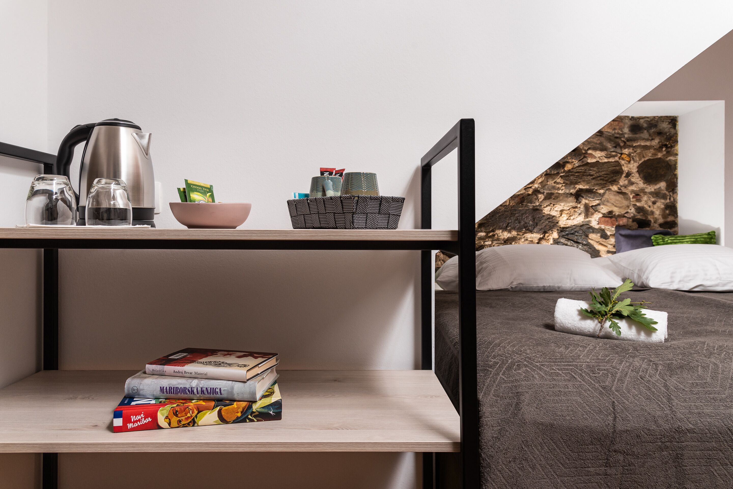 A bedroom with a bed, a shelf with books, a kettle, and a bowl of fruit.