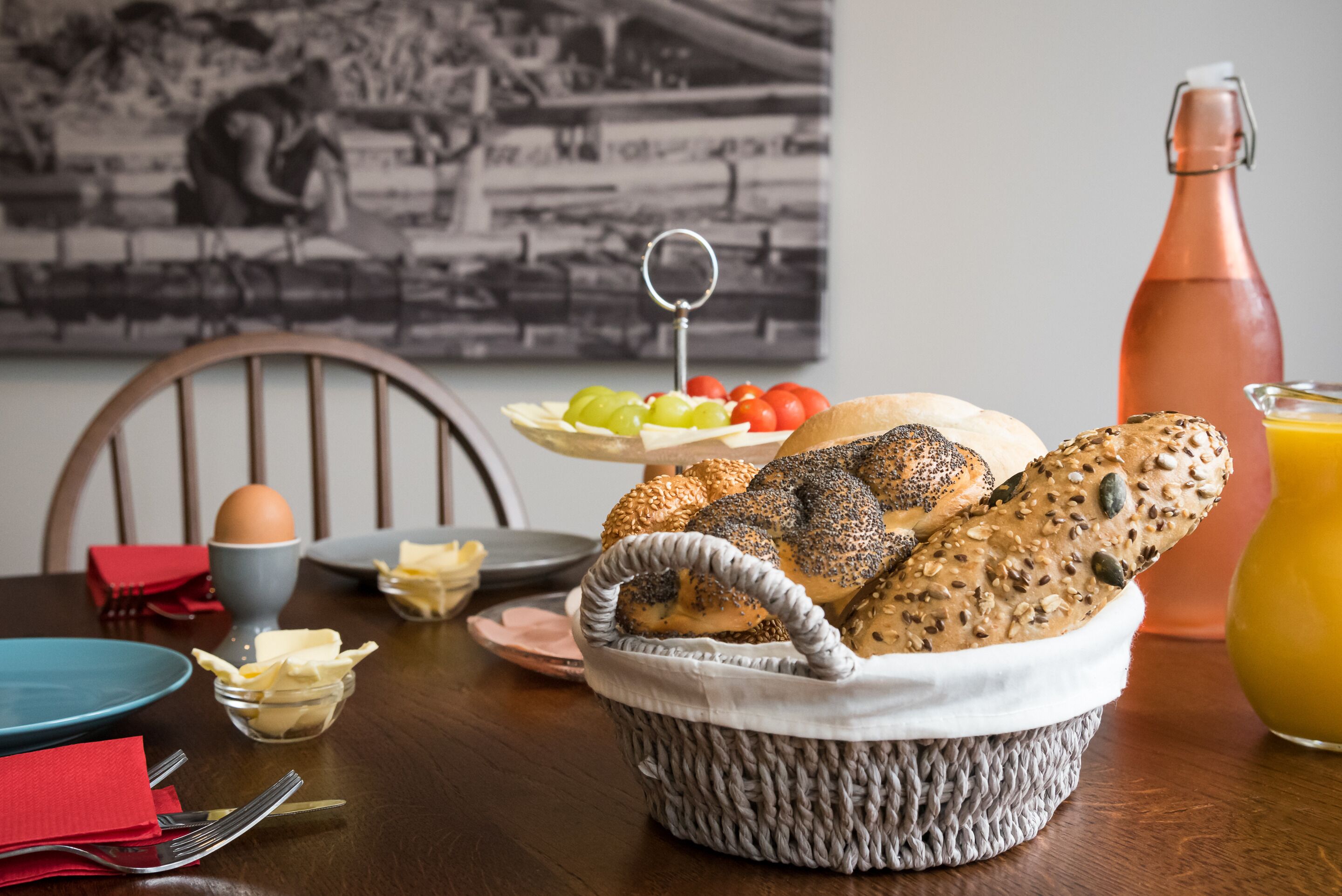 A dining table with a basket of assorted bread, a plate of fruit, a bottle of juice, and a bowl of butter.