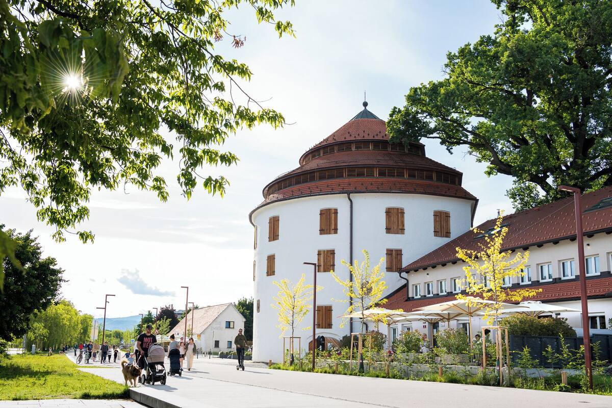 A round building with a red-tiled roof and white walls, surrounded by greenery and a paved walkway.