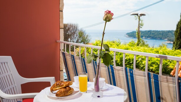 Ein Balkon mit einem Frühstückstisch, komplett mit Croissants und Orangensaft, mit Blick auf eine schöne Aussicht auf das Meer und viel Grün.