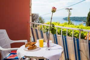 Un balcon avec une table préparée pour le petit-déjeuner, comprenant des croissants et du jus d’orange, offrant une vue magnifique sur la mer et la verdure.