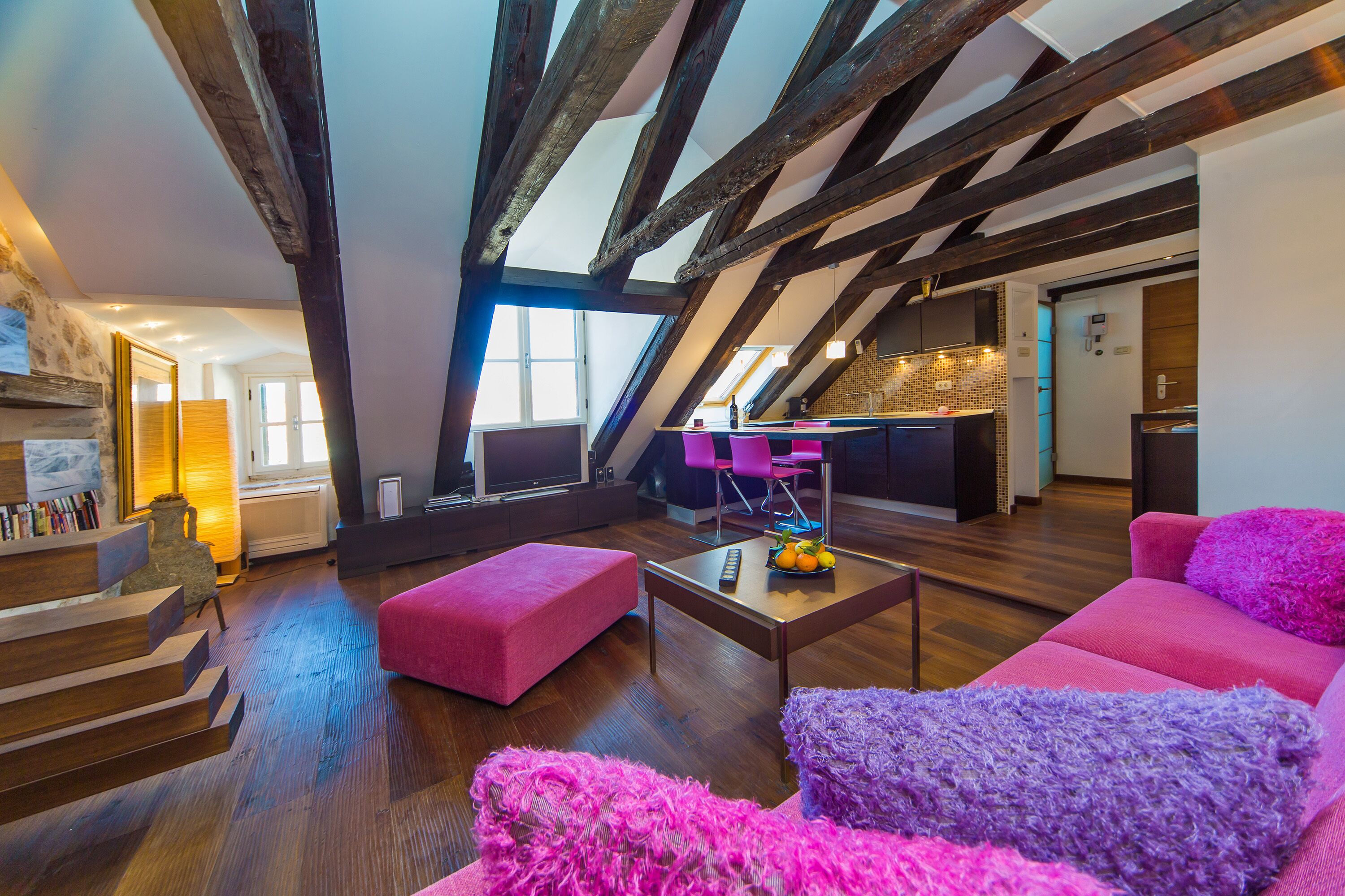 Un loft moderno con vigas de madera a la vista, una zona de estar acogedora con mobiliario rosa y morado vibrante, y una cocina al fondo.