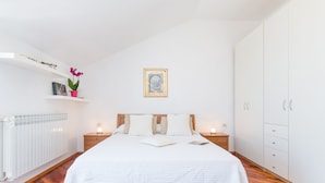A bedroom with a wooden bed, white bedding, a nightstand with a lamp, a radiator, and a white wardrobe. - Villa Mia (Dubrovnik)