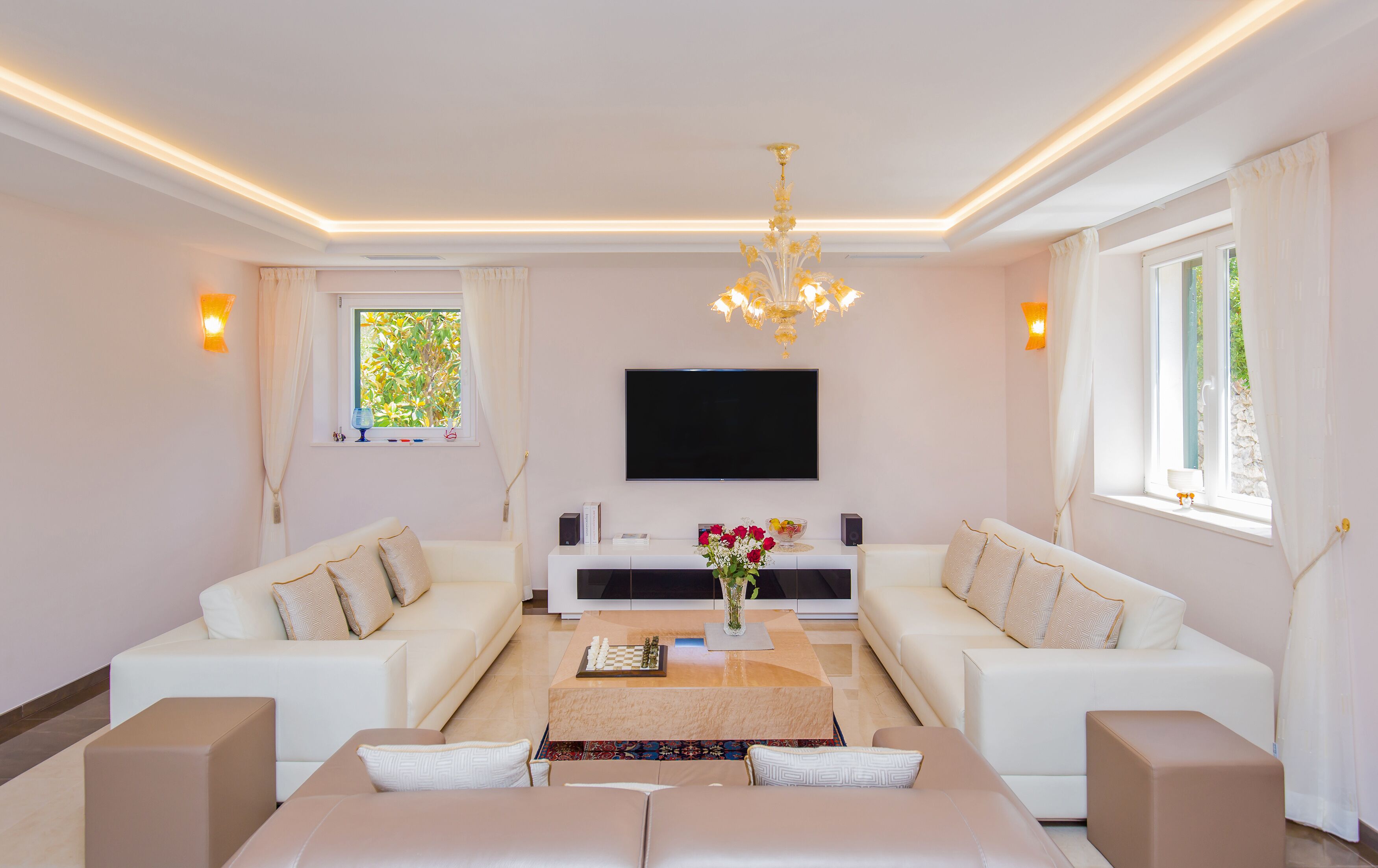 Ein modernes Wohnzimmer mit zwei weißen Sofas, einem Couchtisch, einer Kristallleuchtre, einem Flachbildfernseher und einem Fenster mit Vorhängen.