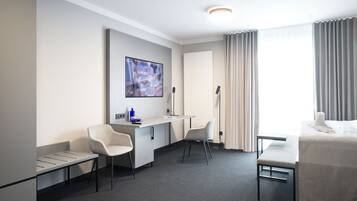 A hotel room with a bed, a desk, two chairs, a flat-screen TV, and a window with curtains.