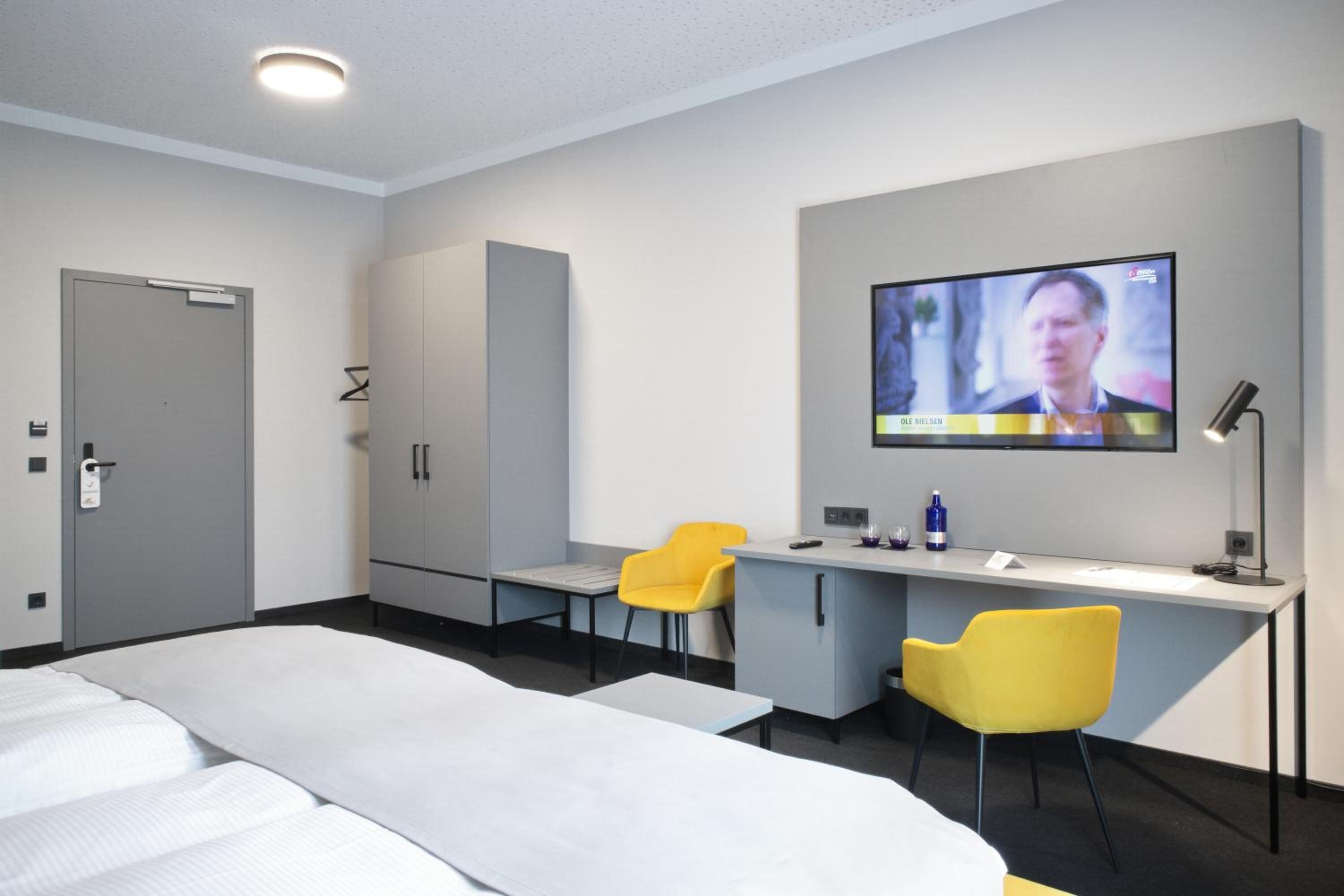 Una habitación de hotel con una cama, un escritorio, dos sillas amarillas, un televisor y un armario.