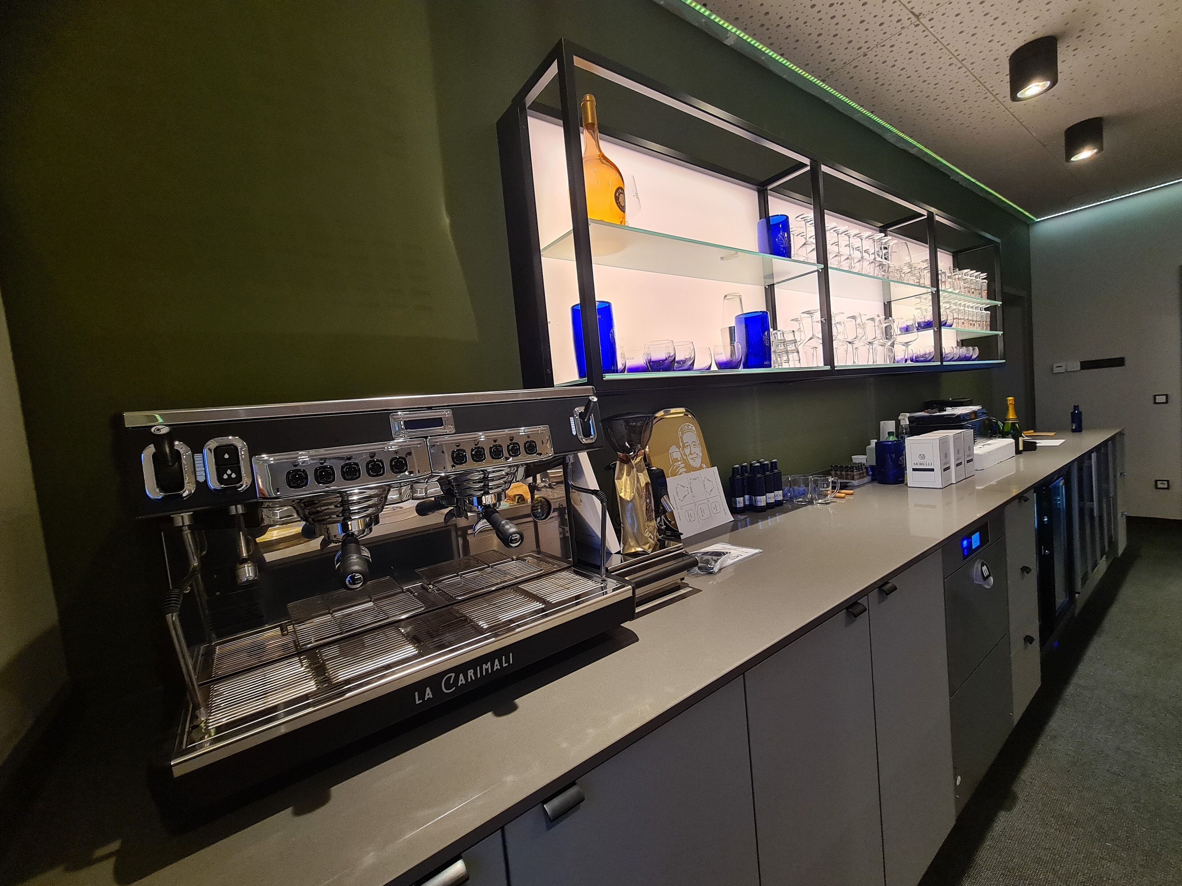 Uma área de bar com uma máquina de café expresso La Carimali, copos e uma seleção de garrafas.