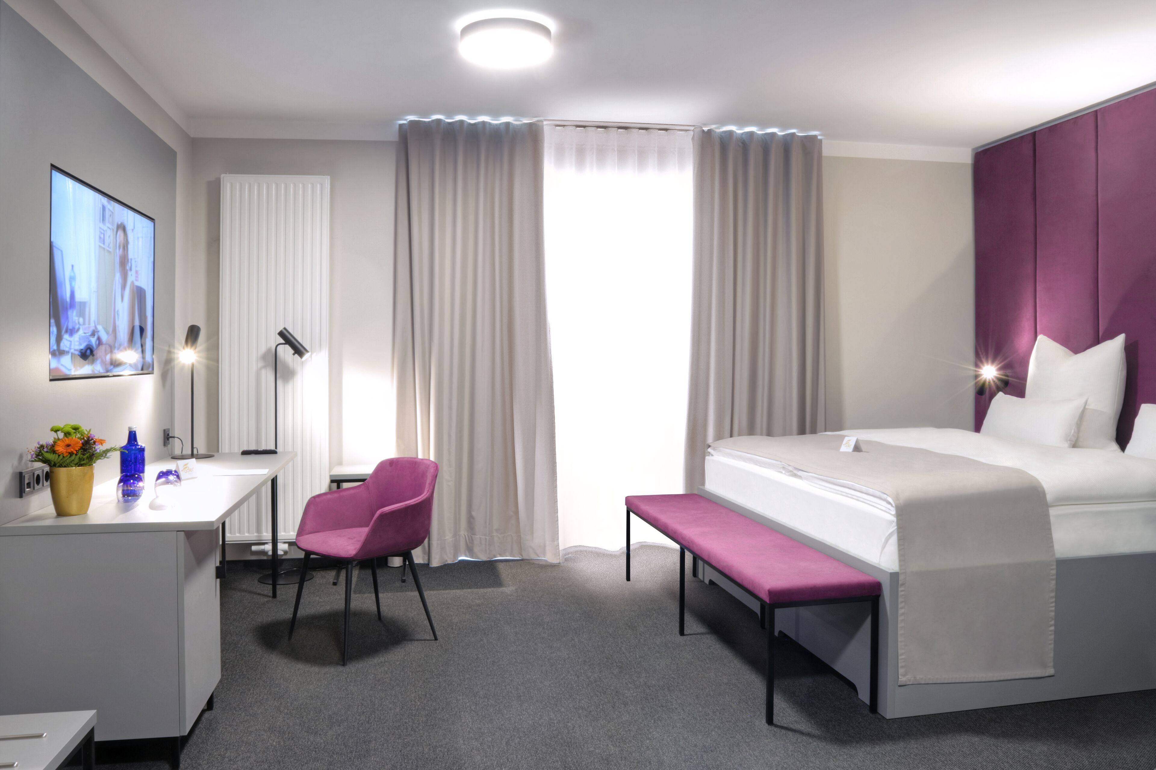 Una habitación de hotel con cama, escritorio, silla, banco, televisión y cortinas.