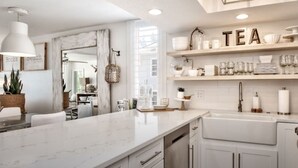 A modern kitchen with white cabinetry, a marble countertop, and a built-in sink. There are shelves above the sink displaying tea-related items.
