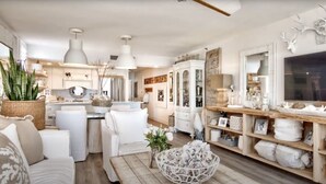 A spacious living room with a large white sofa, a wooden coffee table, and a white armchair. There is a dining area with a wooden table and chairs, and a kitchen area with white cabinets and a large window.