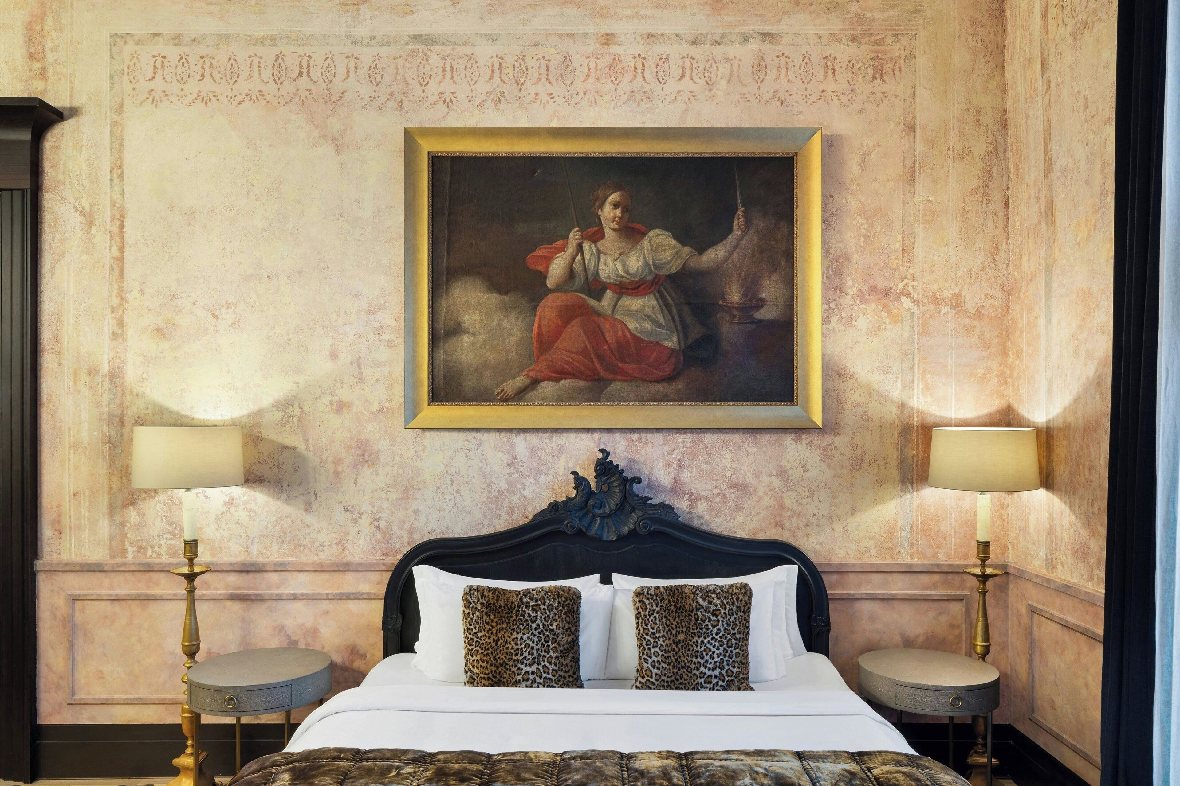 A hotel room with a classic bed, two bedside lamps, and a large framed painting on the wall.