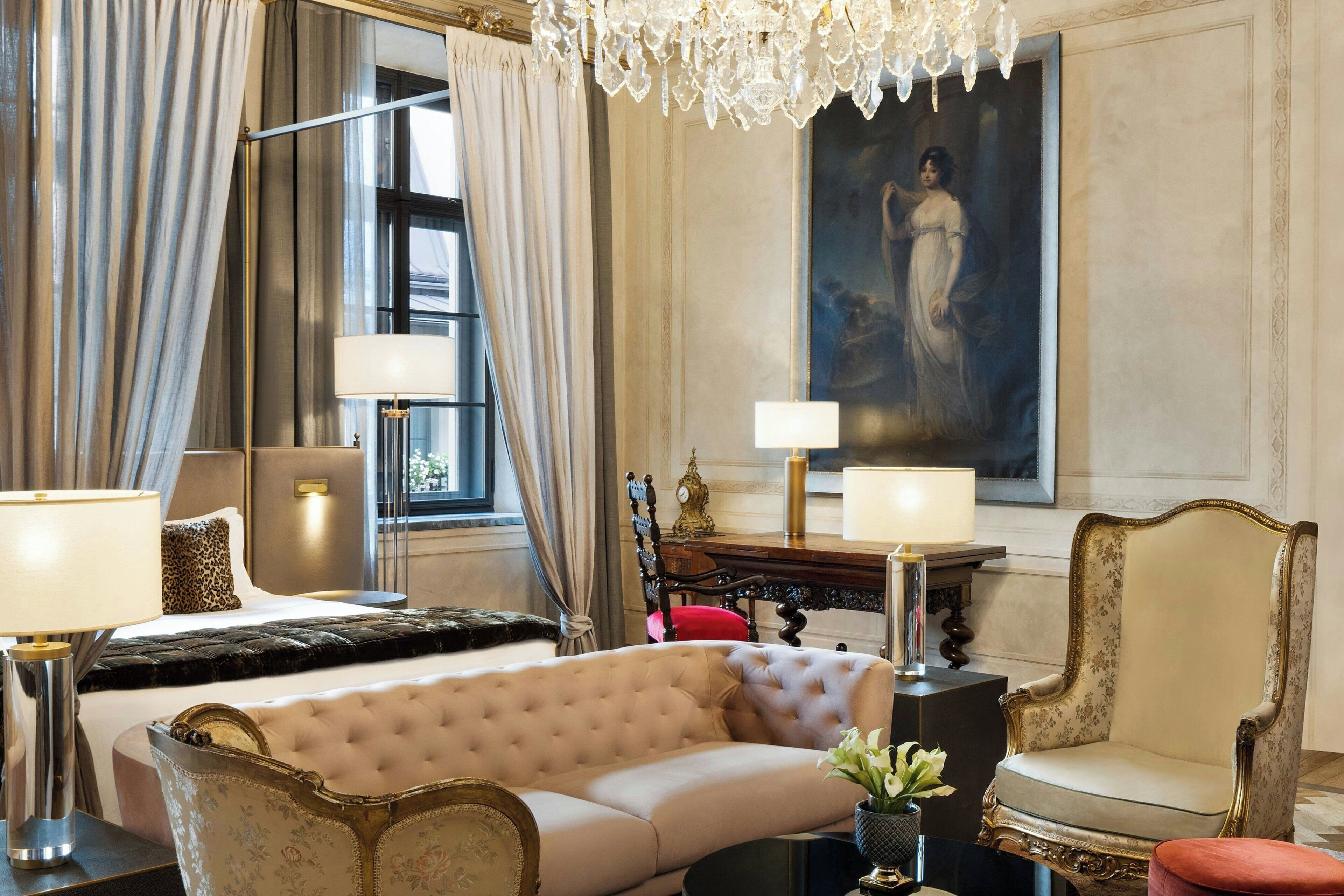 A luxurious hotel room with a large bed, a plush sofa, a classic armchair, a chandelier, and a painting on the wall.