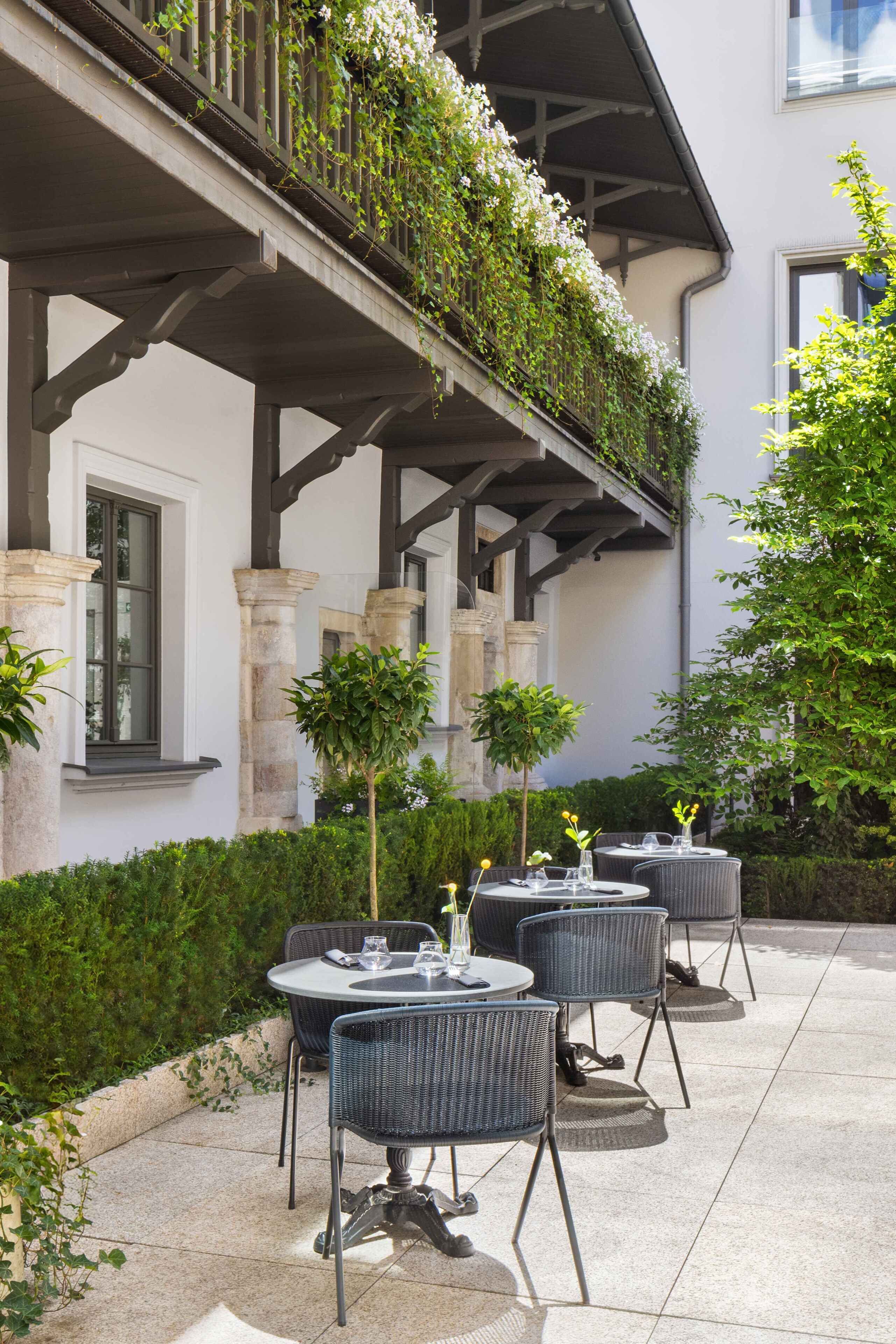 Un patio al aire libre con mesas y sillas redondas, un balcón con plantas trepadoras y un seto.
