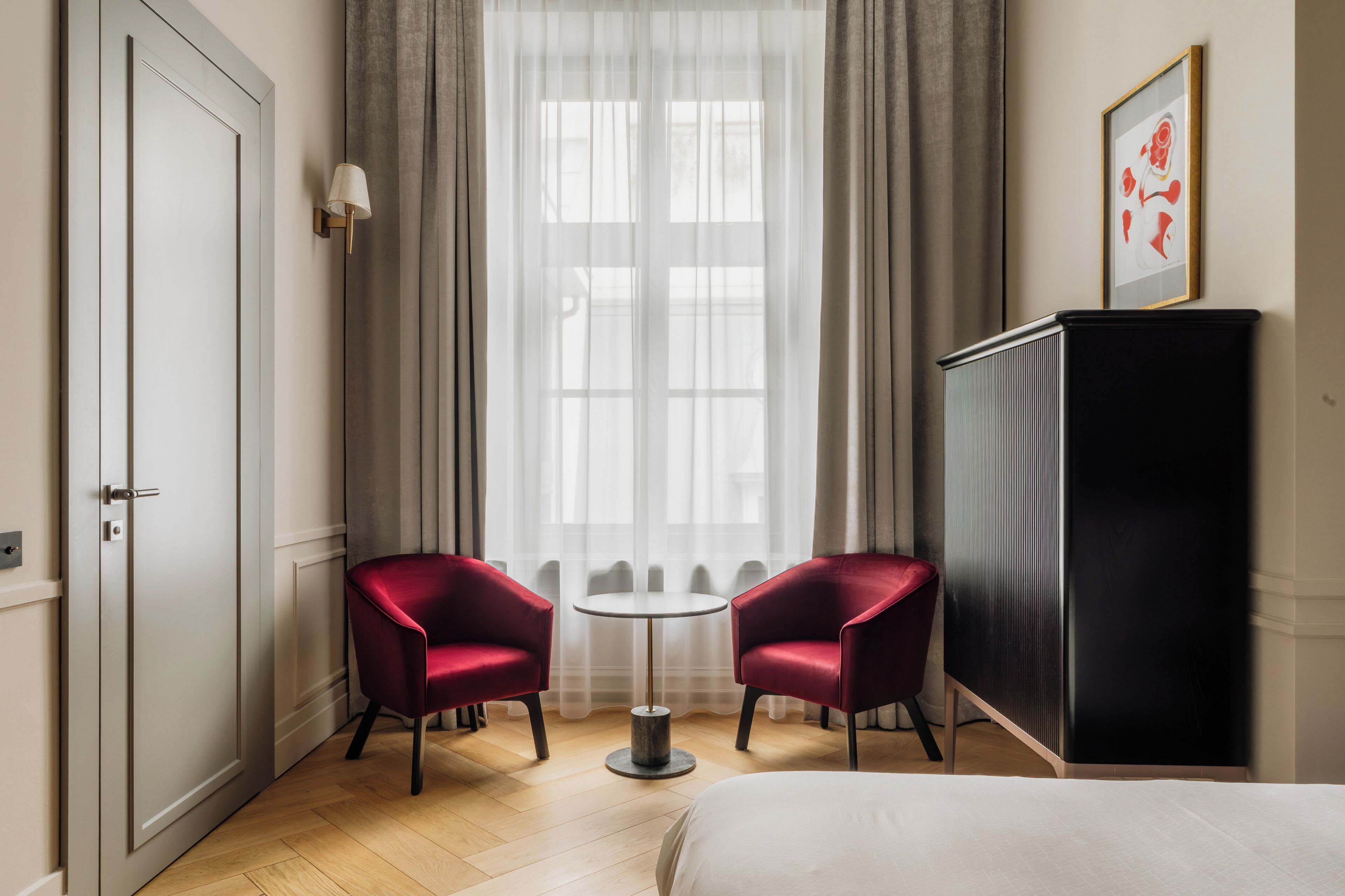 Une chambre d’hôtel avec un lit, deux fauteuils rouges, une petite table ronde, une armoire et une fenêtre avec des rideaux légers.