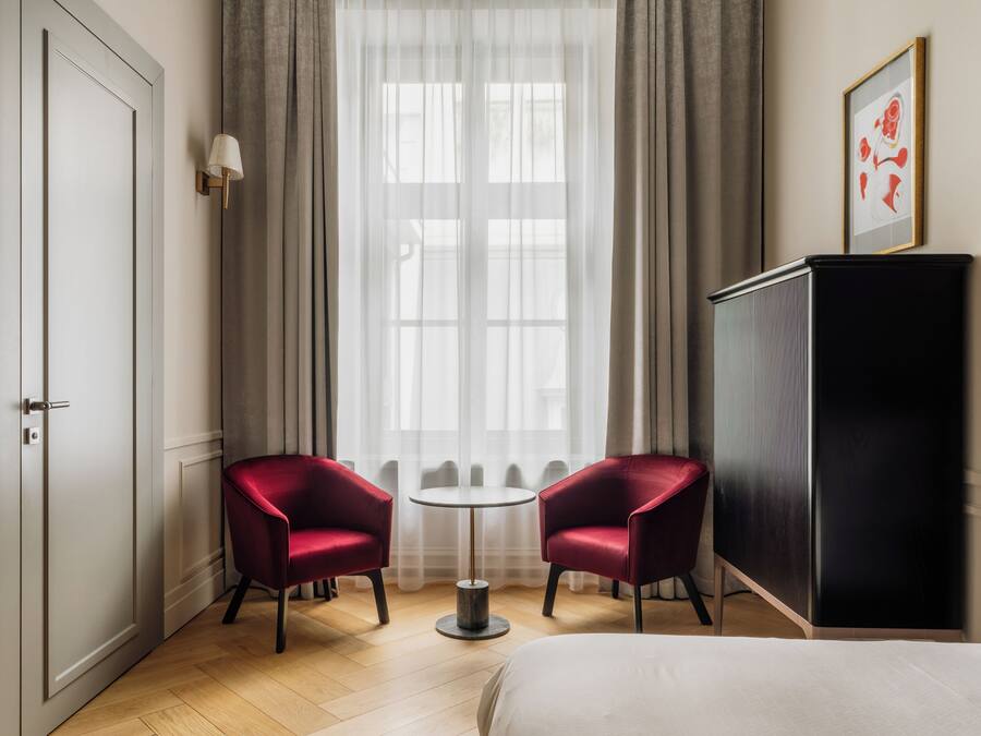 A hotel room with a bed, two red chairs, a small round table, a wardrobe, and a window with sheer curtains.