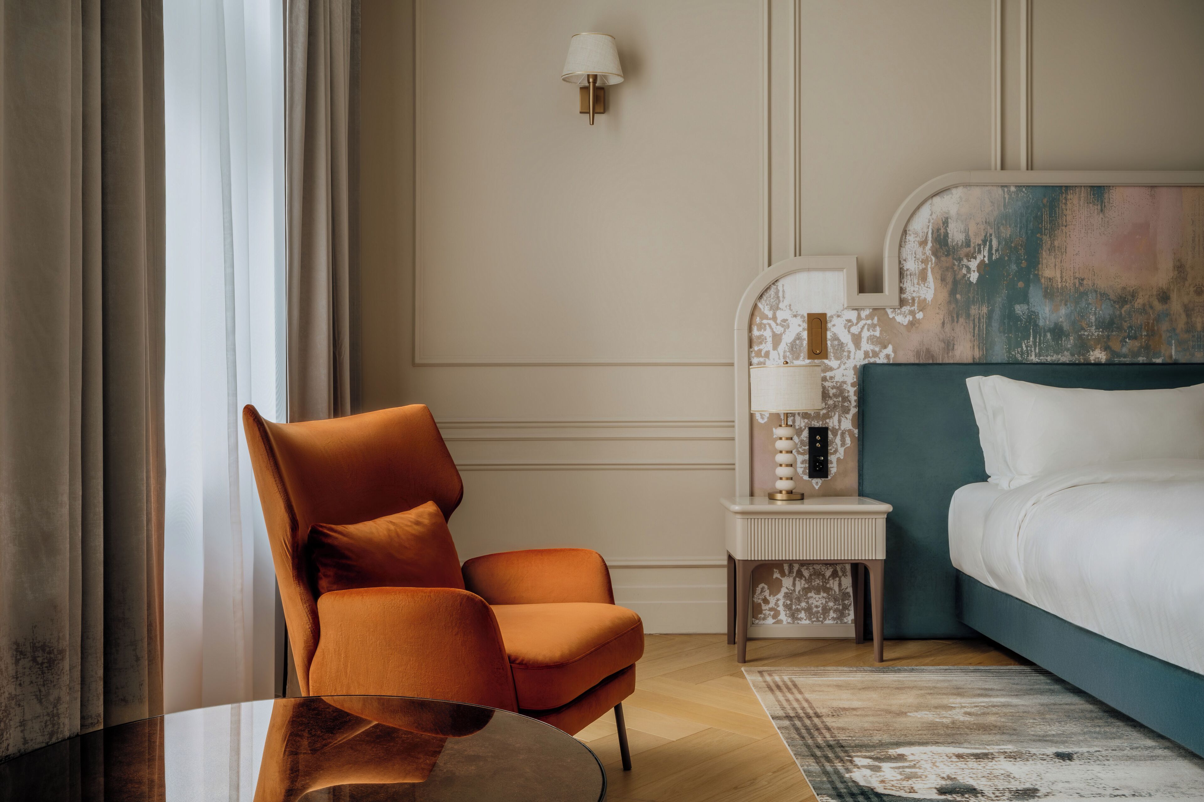 Ein Hotelzimmer mit einem türkisfarbenen Kopfteil, einem Nachttisch aus Holz mit Lampe, einem orangefarbenen Sessel und einem Glastisch.