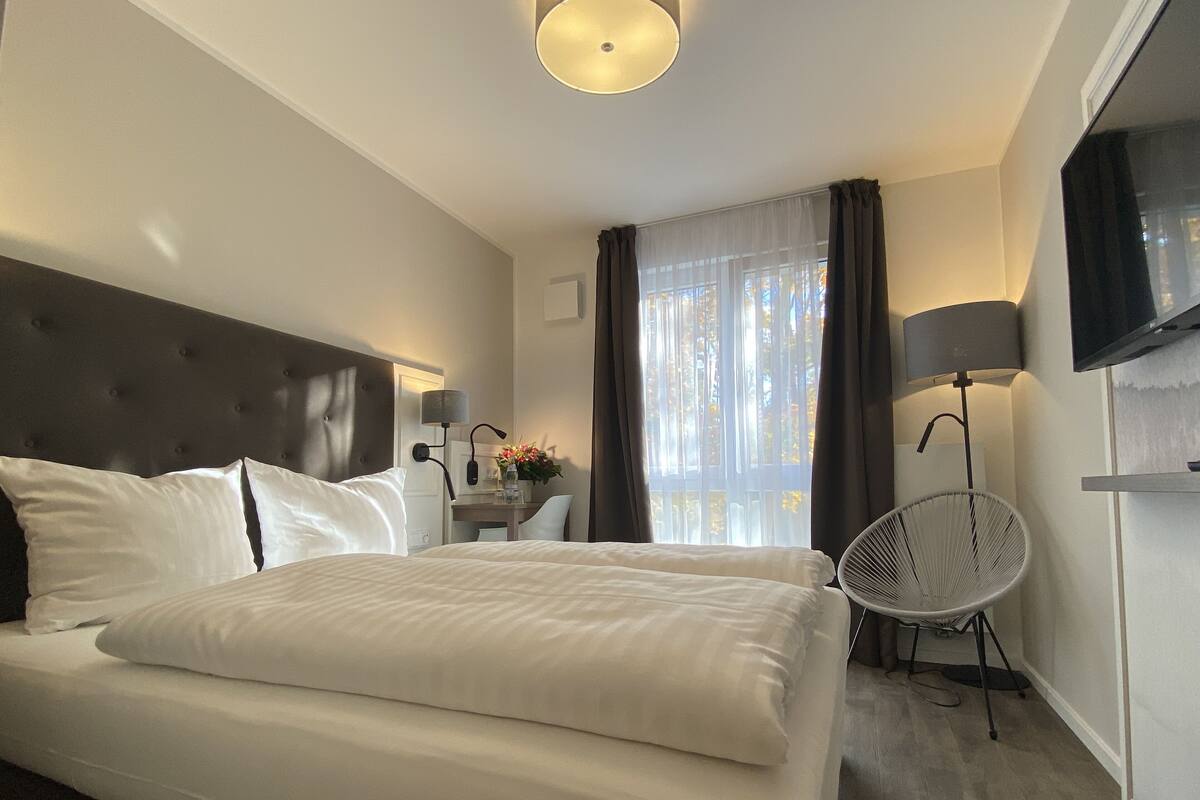A hotel room with a large bed, a desk with a lamp, a chair, a TV, and a window with curtains.