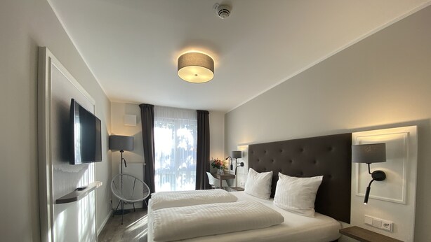 A modern hotel room with a large bed, a flat-screen TV, a desk with a chair, and a window with curtains. - Signature East Hotel Falk (Hamburg)