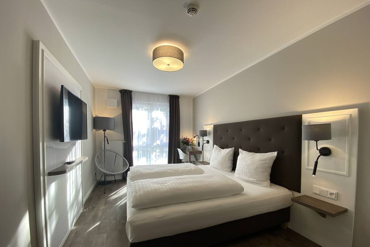 A modern hotel room with a large bed, a flat-screen TV, a desk with a chair, and a window with curtains.