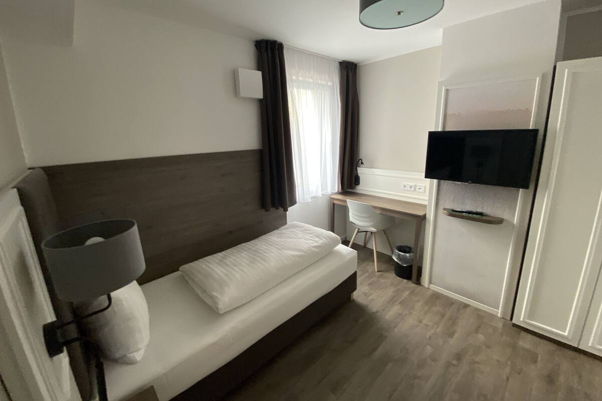 A modern hotel room with a bed, a desk, a chair, a television, and a wardrobe.