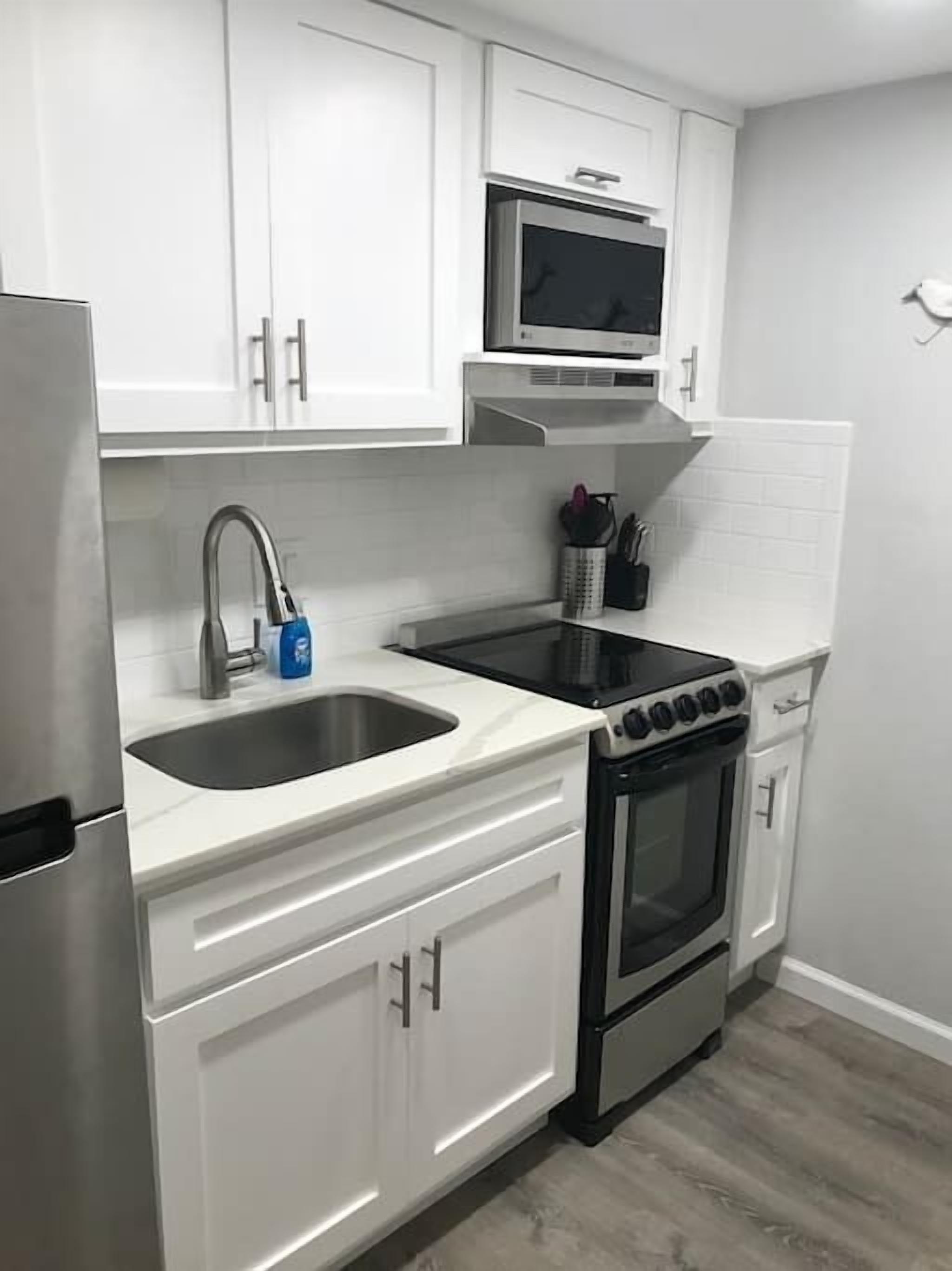 A compact kitchen with white cabinets, a stainless steel refrigerator, a microwave, a stove, and a sink.